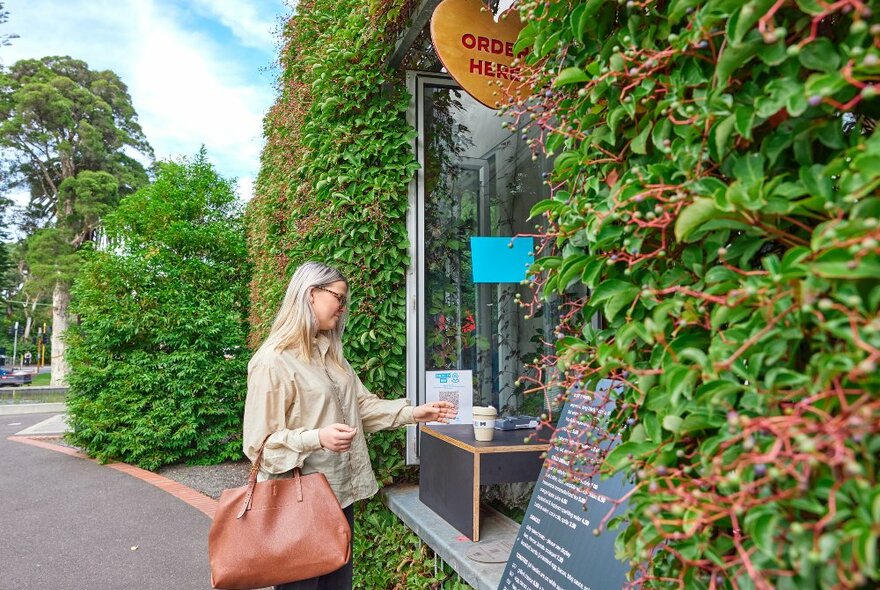 A woman is ordering coffee at a cafe window. The wall is covered in green vines.