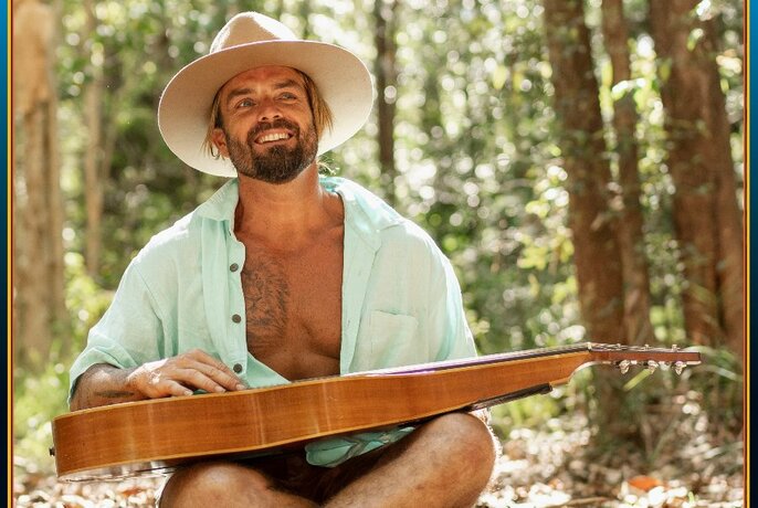 Musician Xavier Rudd looking relaxed in a forest with a guitar on his lap, a wide-brim hat and his shirt unbuttoned.