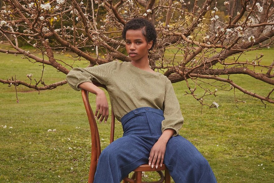A model leaning back in a chair outside wearing high waisted blue pants and a khaki coloured long sleeve loose top.