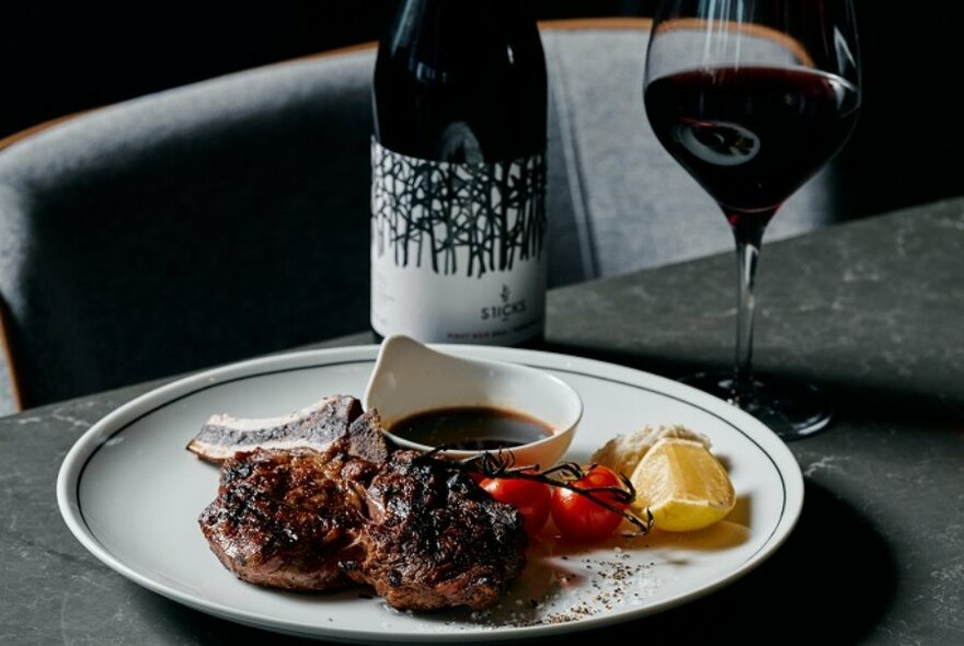 Grilled meat arranged on a white plate with sides and a sauce, a glass of red wine and a wine bottle.