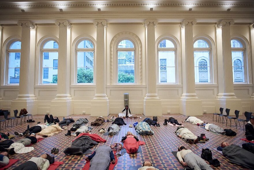 Many people laying on mats on the floor of a large old building with arched windows.