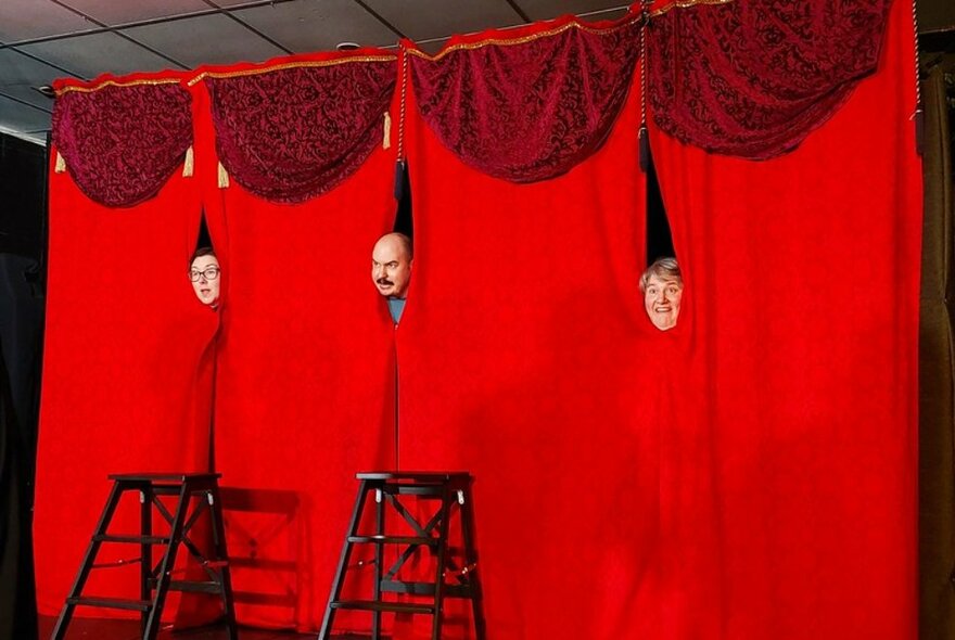 Three faces poking through gaps in a long red curtin on a stage with stools.