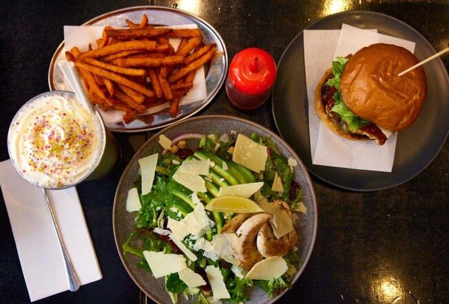 A burger on a plate next to a chicken salad, a plate of sweet potato fries and a milkshake.