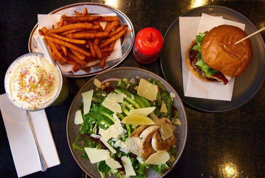 A burger on a plate next to a chicken salad, a plate of sweet potato fries and a milkshake.