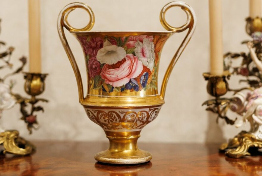 An ornate golden vase with two curled handles and handpainted floral blooms on the main bowl.