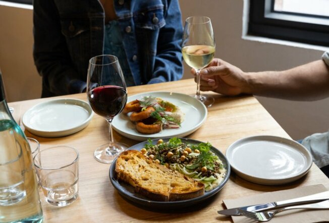 Close up shot of two plates of food with white and red wine. Two people are sat at the table about to eat.