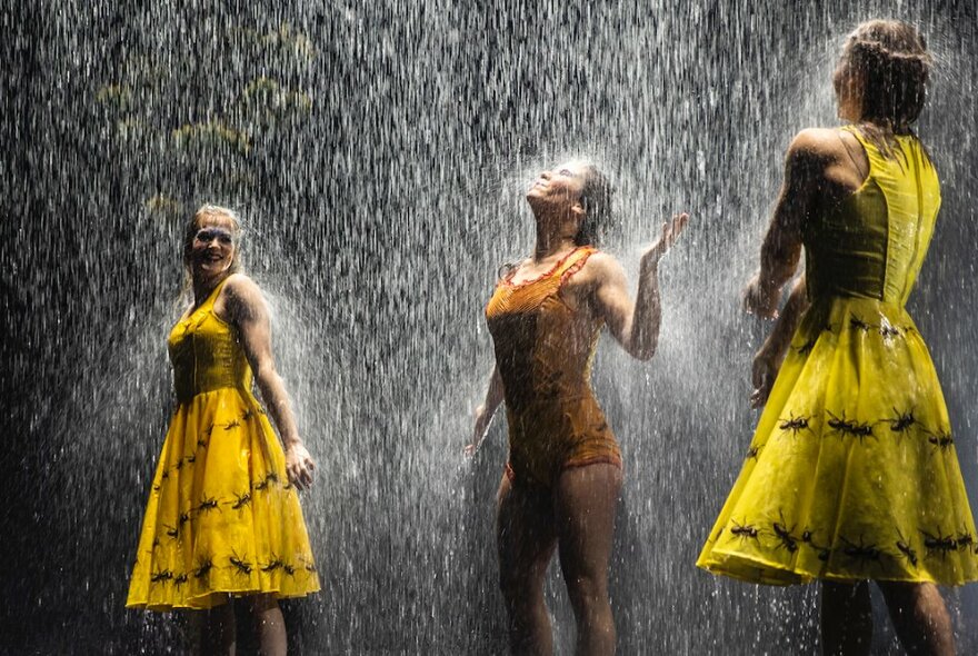 Performers wearing yellow and orange outfits standing on stage under a downpour of rain.