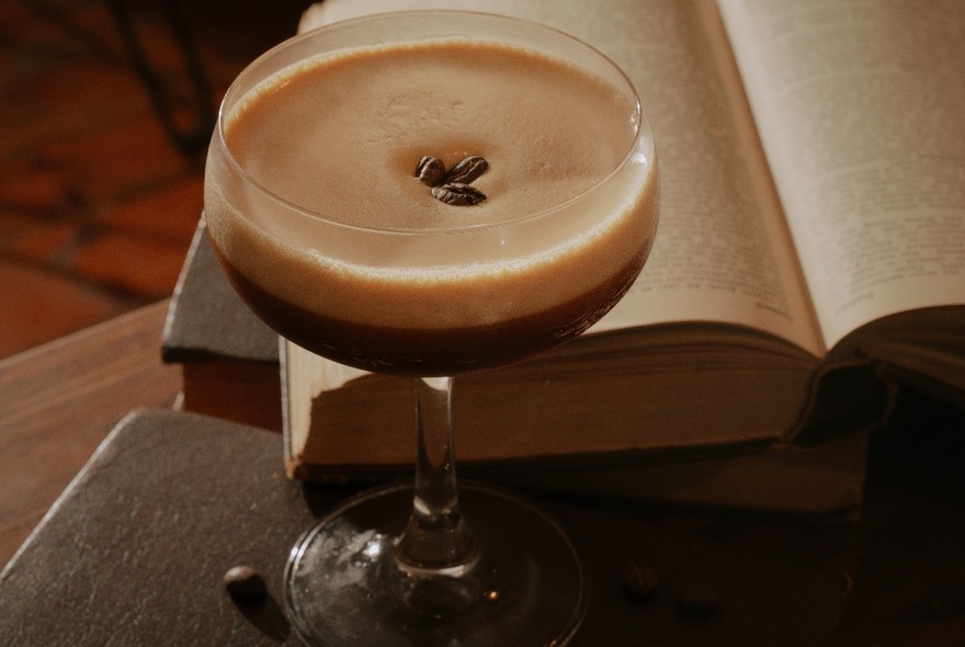 A creamy espresso martini cocktail, with three coffee beans as garnish, an open book in the background.