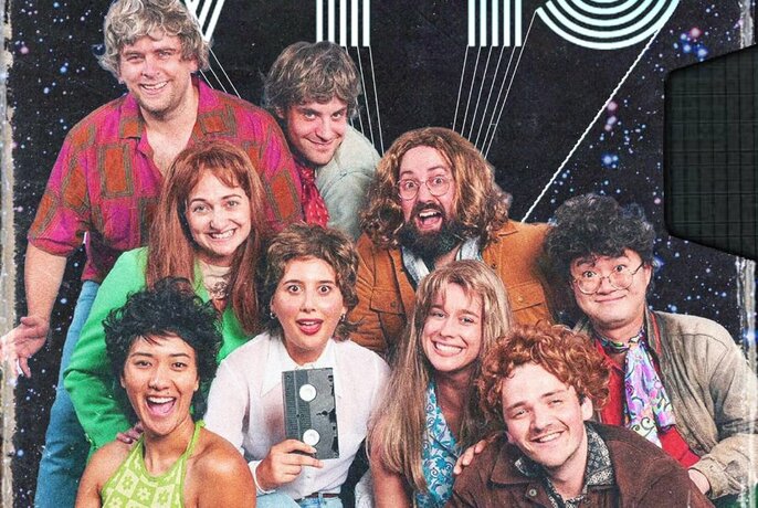 Group of actors in 1970s inspired outfits and wigs smiling and posing.