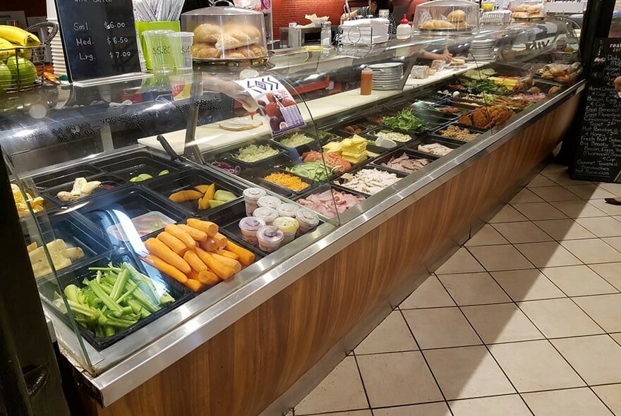 Glass cafe food counter filled with vegetables and trays of sandwich ingredients.