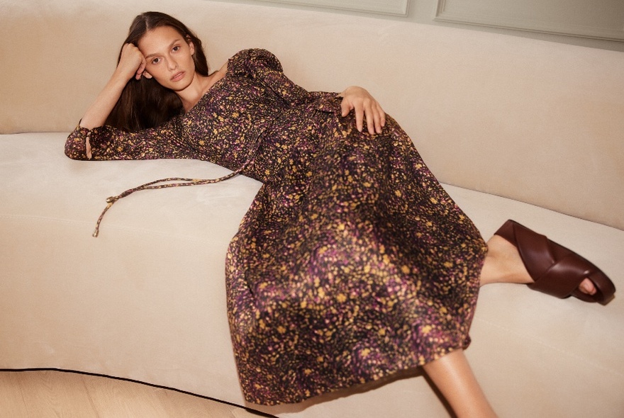 Model lounging on a beige sofa wearing a calf-length bronze patterned dress.