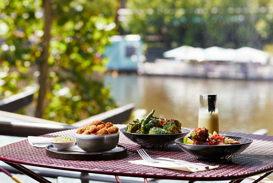 An outdoor restaurant table beside the river, set with three dishes and a cocktail.