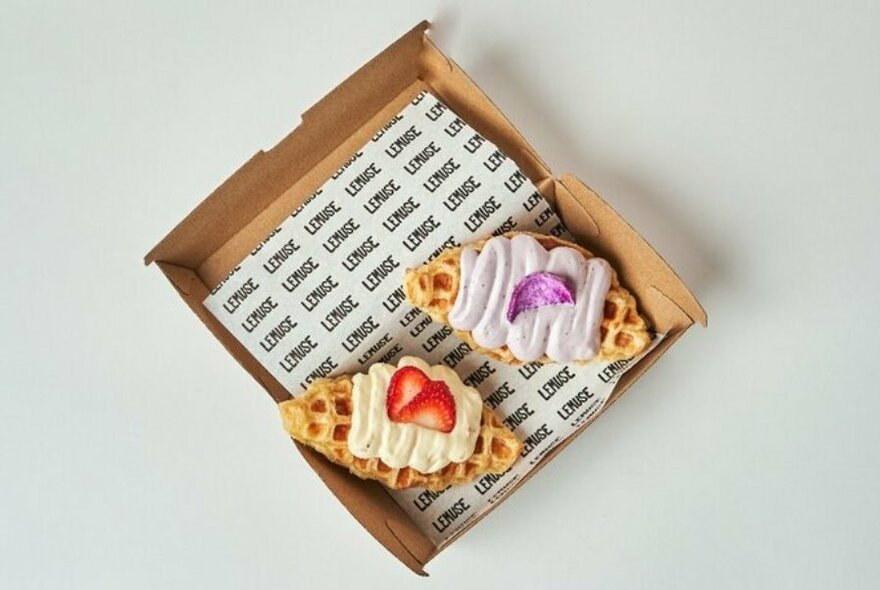 Two sweet pastries, with a cream and fruit garnish, resting in an open cardboard takeaway container. 