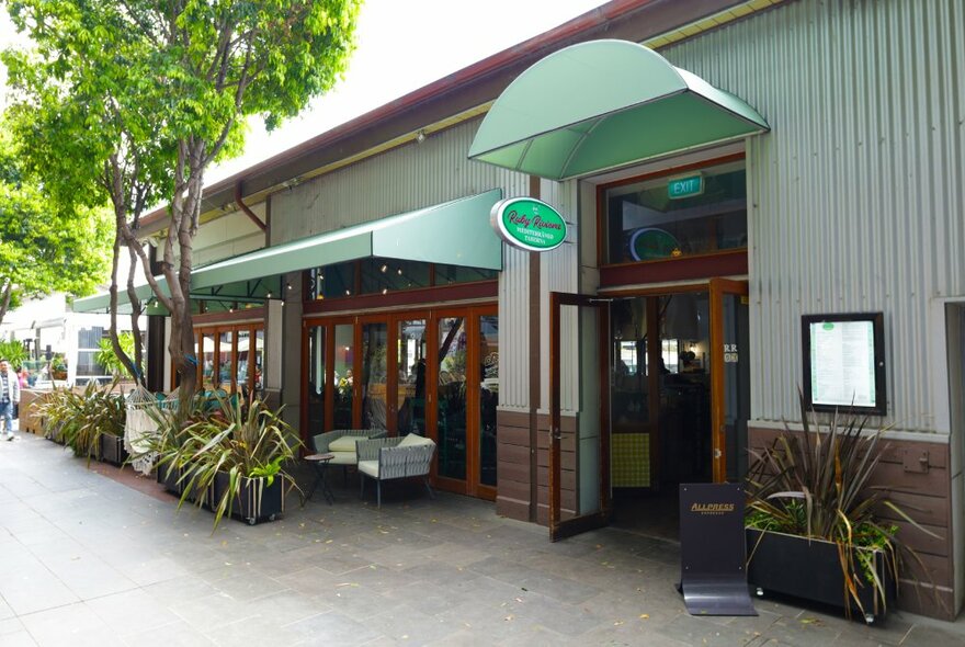The outside of an Italian taverna with grey corrugated iron walls, pale green awnings and a tree. 