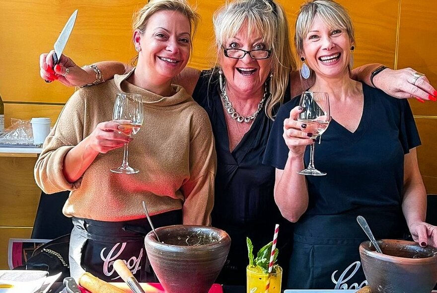 Three women at a Thai cooking workshop with wine, and one holding a chef's knife.
