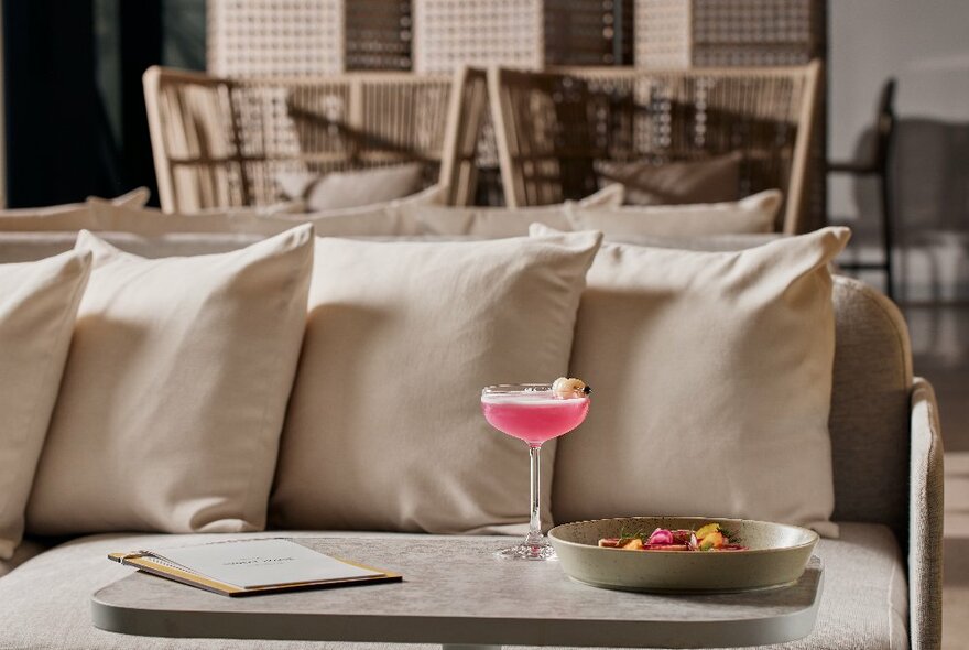 A pink cocktail and dish of food on a table in frton of a beige cushioned sofa.