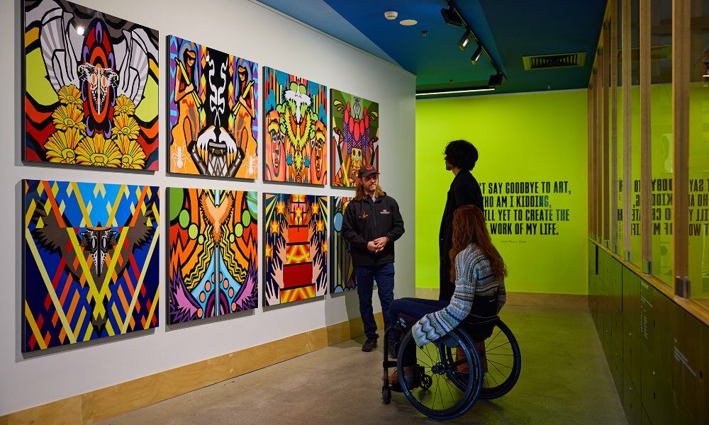 A guide in a gallery showing a series of colourful paintings to two people. 