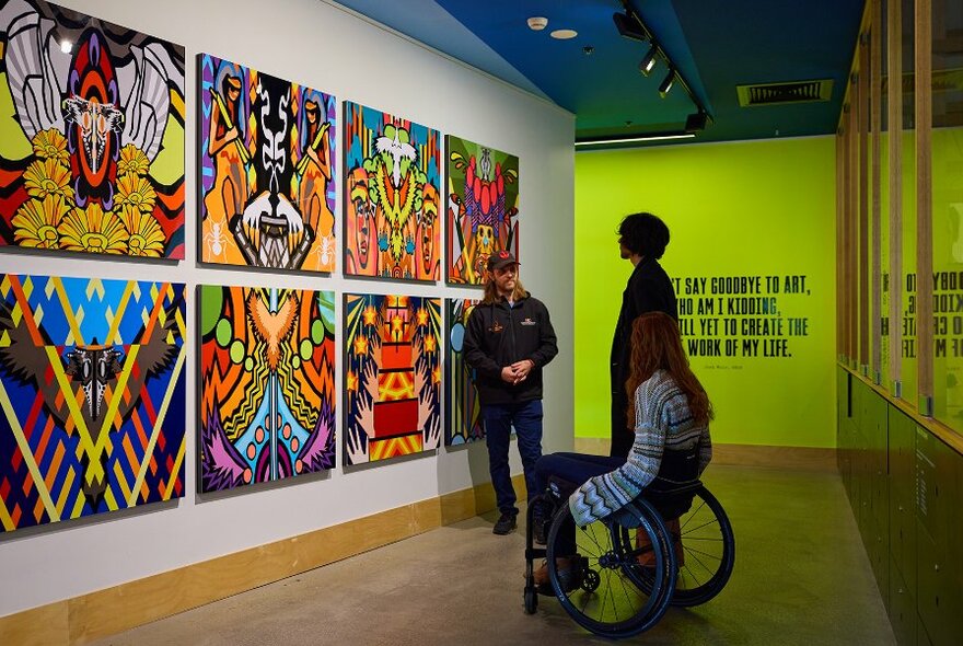 A guide in a gallery showing a series of colourful paintings to two people. 