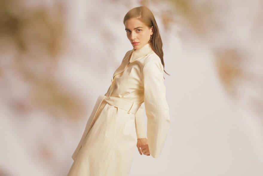 A model wearing a cream coloured long sleeved belted coat, leaning back slightly in profile against a blurred background. 