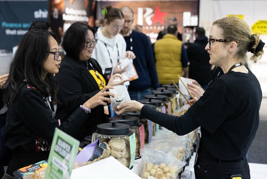 Women at a stall at the Melbourne International Coffee Expo being offered a taste test in a small container.