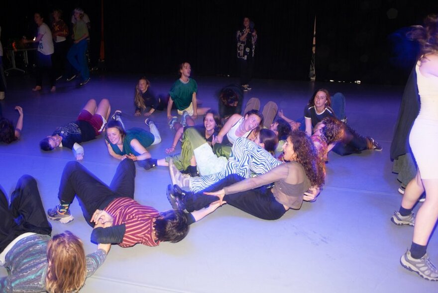 A group of people arm in arm whilst either lying on the blue floor or falling down.