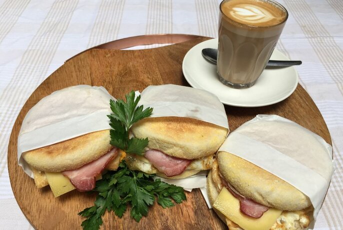 Wooden board with three toasted sandwiches in paper, coffee in glass at rear.