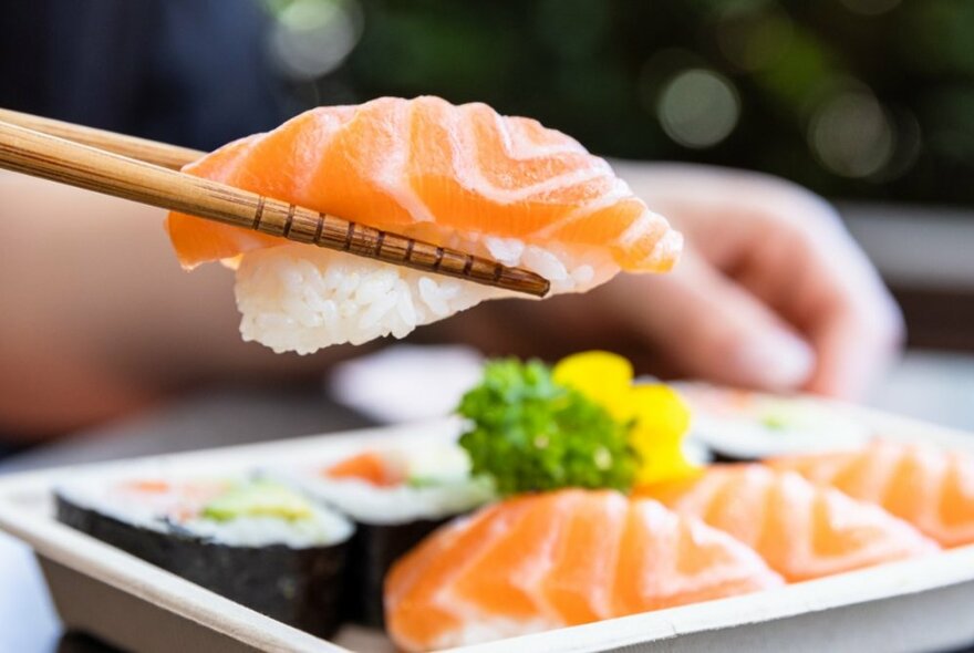 Chopsticks holding a piece of salmon sushi above a dish of Japanese food.