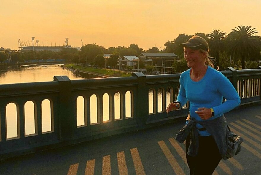 A jogger running across a bridge at sunrise with the MCG in the background.