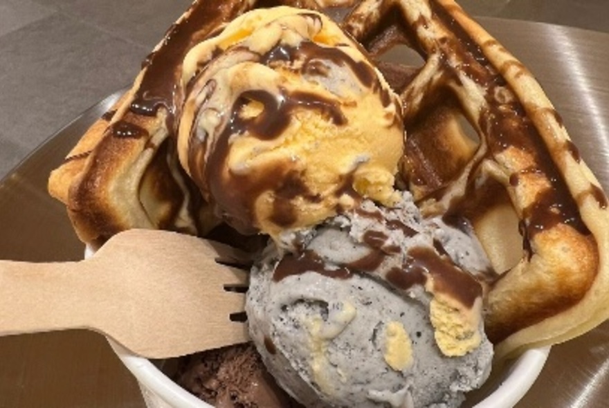 Black sesame and mango flavoured ice cream scoops in a white paper cup with waffles, drizzled with chocolate sauce; a bamboo fork. 
