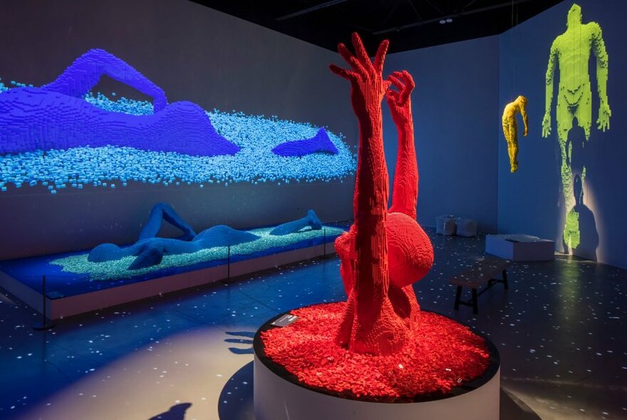 Sculpture of a half torso with arms raised above its head, crafted from many red lego bricks, and a sculpture of a person swimming, made from blue lego bricks, on display in a large exhibition space.