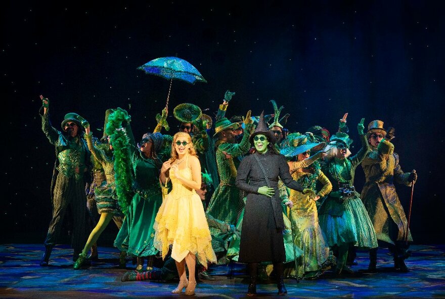 The cast of WICKED the Musical, onstage in costume, with Glinda the 'Good' Witch, and Elphaba the 'Evil' Witch at the front.