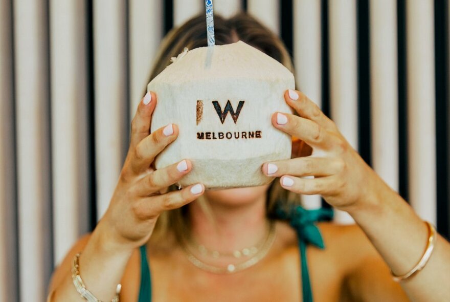 A person holds up a coconut with a straw in front of their face with the words W Melbourne stamped on it. 