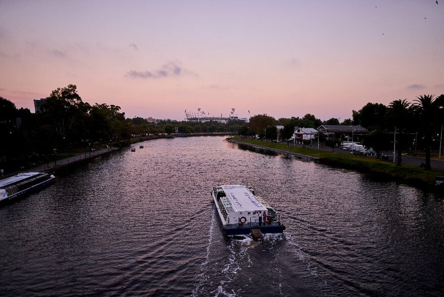 A riverboat cruising up the Yarra River at dusk, with the MCG on the horizon.