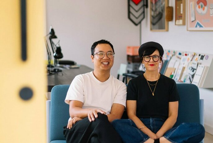 Designers Daniel and Emma sitting next to each other on a couch in a studio environment, artwork and a table visible in the background.