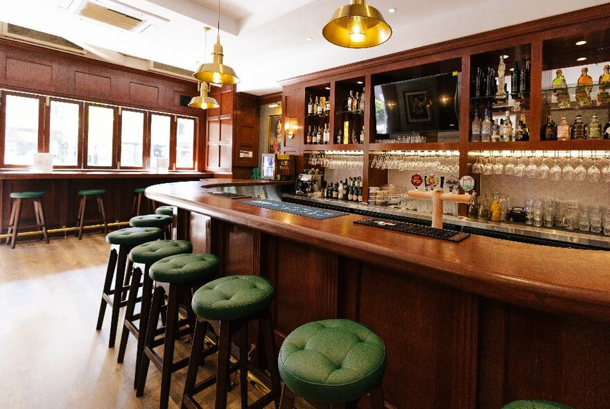 The interior of a traditional looking pub with a curved bar and green upholstered stools. 