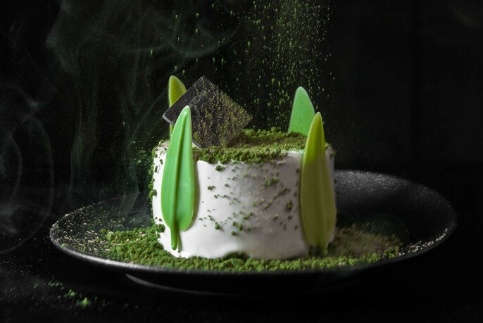 White, round dessert with icing green leaves on the side scattered with green powder.