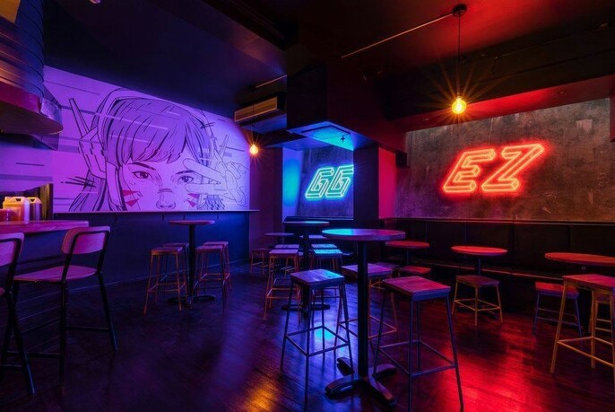 Darkly lit GG EZ interior with stools and tables, large purple screens and neon lights.