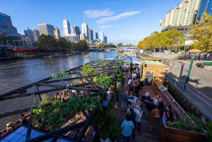People drinking on the riverside Arbory Bar moored on the Yarra River with city buildings in the background.