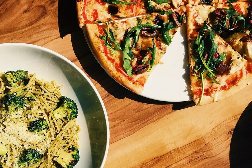 A pizza and pasta dish on a wooden table.
