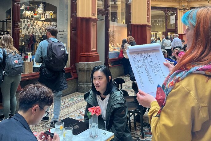 A person holding a sketch while looking at people seated at a cafe in a Melbourne arcade.