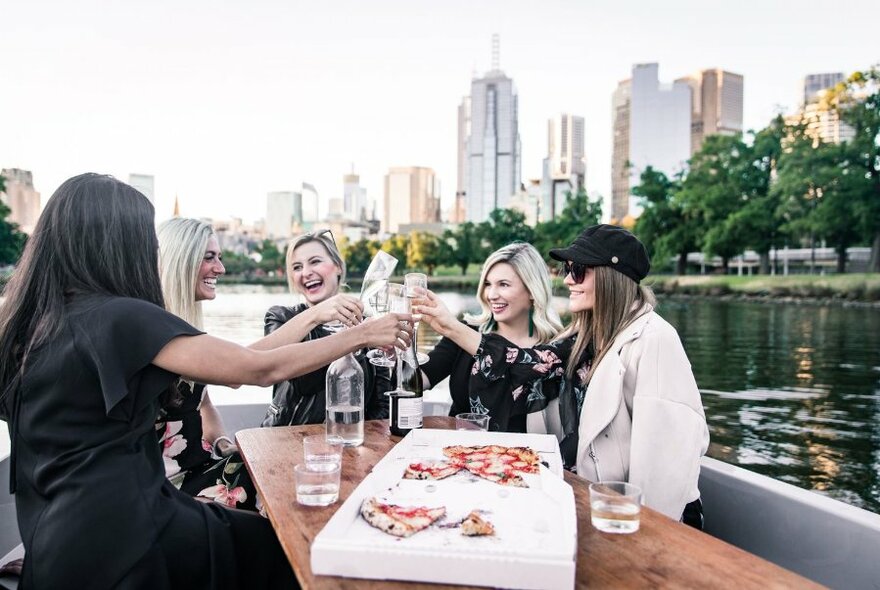 A group of women on a boat toasting with champagne glasses with a pizza box on the table. 