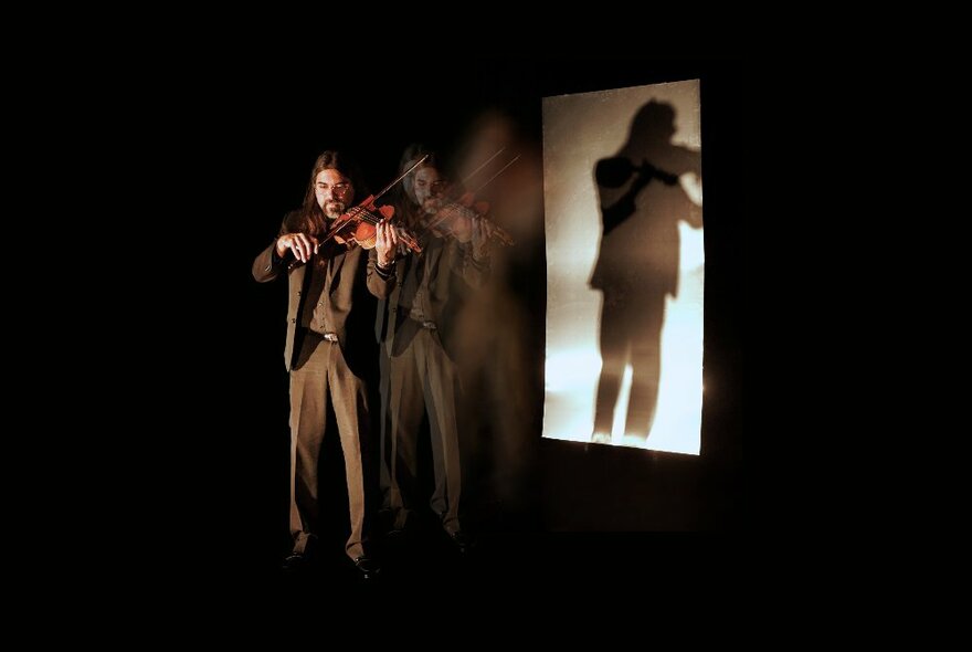 A man playing a viola in front of a projection screen on a dark stage, his reflection visible on the screen behind. 