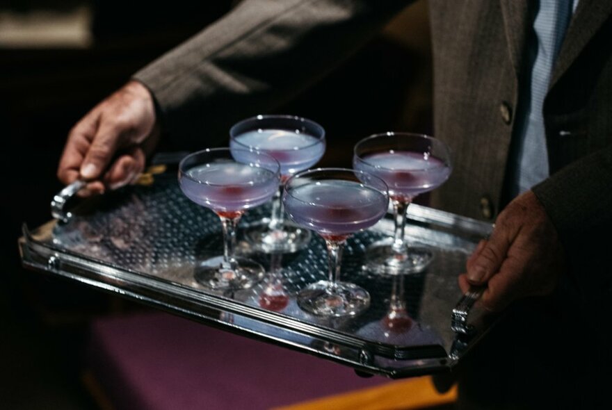 Four lavender coloured cocktails in classic tulip shaped glasses, being carried on a silver tray.