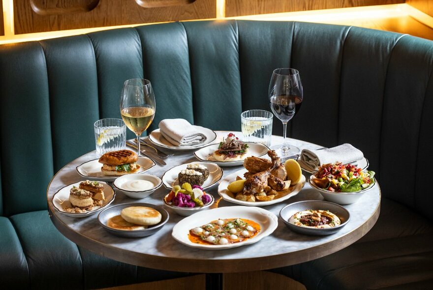 A table in a restaurant with plates of food, glasses of wine, and napkins, and a rounded banquette seat behind it.