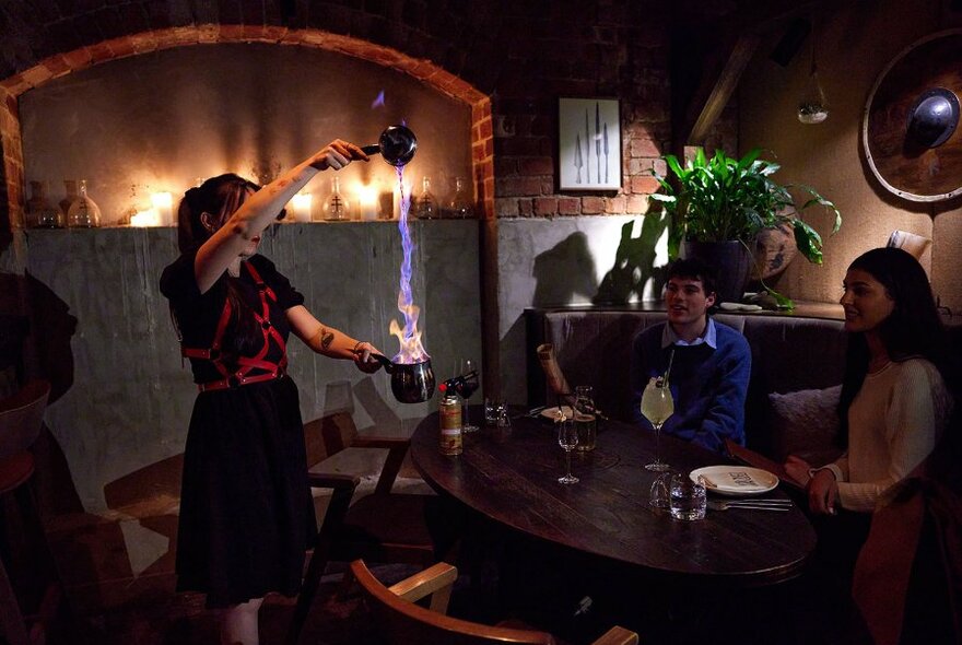 A bartender pouring a flaming cocktail as a couple watch in a dimly lit bar. 