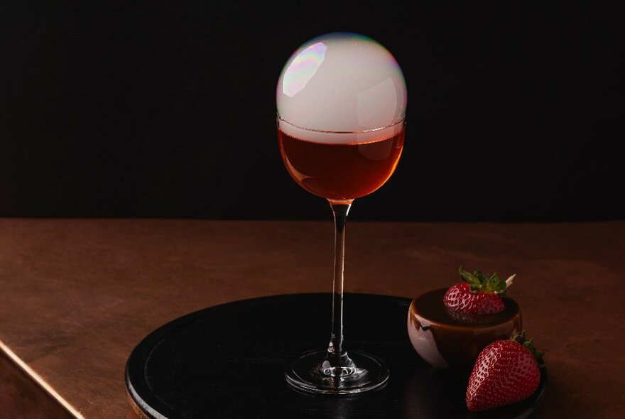 A red cocktail with a cloudy dome, on a black tray next to two strawberries.