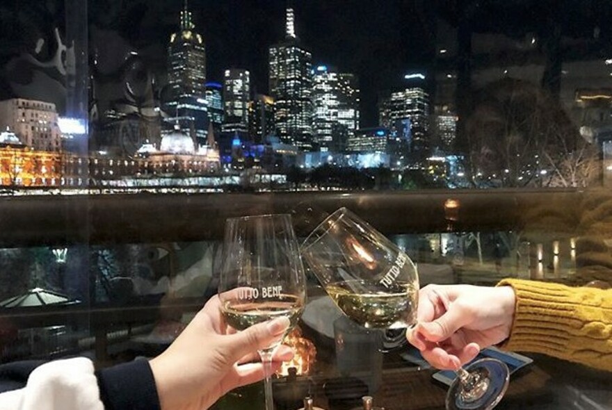 Two outstretched arms clinking glasses with the Melbourne skyline at night in the background. 
