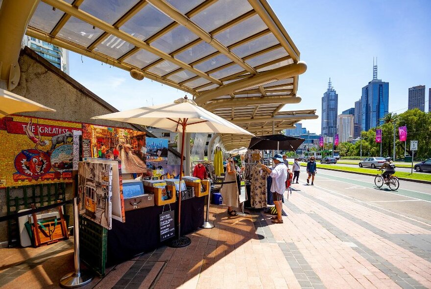 Outdoor view of market stalls along the Arts Centre Promenade with St Kilda Road and city visible in the background.
