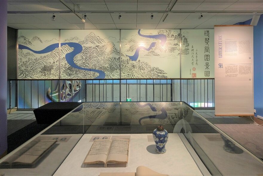 Exhibits on display in a glass display case in a room with a large Chinese illustration of a map on the wall in the background.