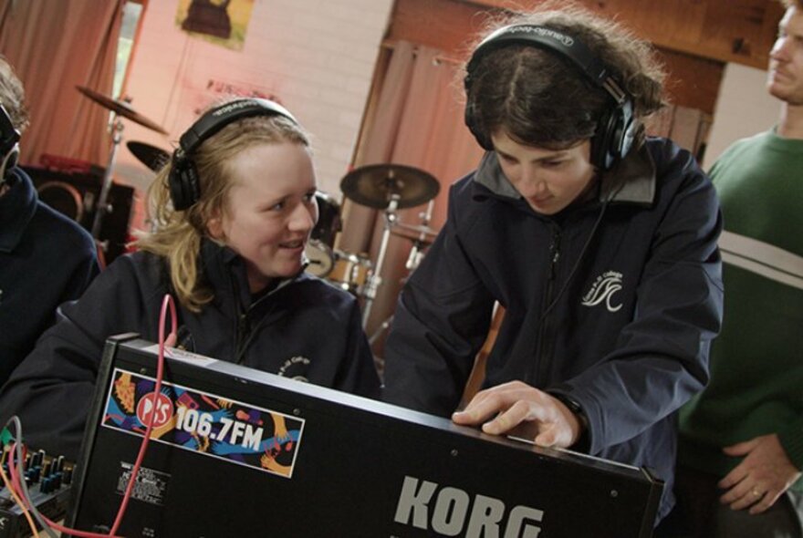 Two people seated at a Korg synth wearing headphones.